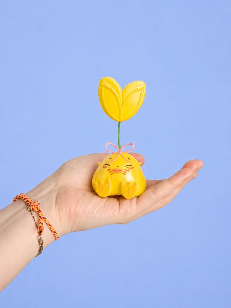 A Humulus figure with a yellow tulip and a pink bow, held in an open hand